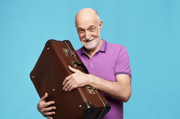 Koncepcja ludzie, starzenie się, podróże i turystyka. Atrakcyjny radosny podekscytowany starszy mężczyzna w fioletowej koszulce, który zamierza spędzić wakacje w tropikalnym kraju, uśmiechając się szeroko, trzymając skórzaną torbę bagażową