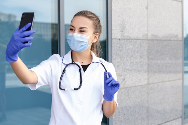 Koncepcja medycyny, opieki zdrowotnej, zawodu i ludzi - europejska lekarka ze stetoskopem w sukni medycznej, gumowych rękawiczkach i masce ochronnej, robiąca selfie na swoim telefonie z wolną przestrzenią