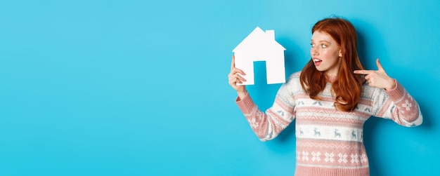 Koncepcja nieruchomości podekscytowana rudowłosa kobieta z rudymi włosami wskazująca i patrząca na model domu z papieru