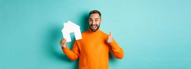 Bezpłatne zdjęcie koncepcja nieruchomości zadowolony człowiek polecający agencję pokazującą kciuk w górę i dom maket stojący nad
