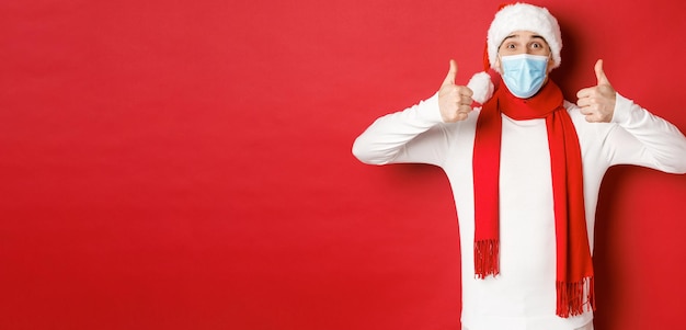 Koncepcja nowego roku, koronawirusa i świąt. wesoły mężczyzna świętujący nowy rok i zdystansowanie społeczne, ubrany w maskę medyczną, czapkę mikołaja i szalik, pokazując kciuk w górę z aprobatą.