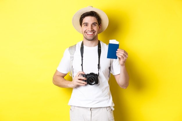 Koncepcja podróży, wakacji i turystyki. Uśmiechnięty mężczyzna turystyczny trzymając aparat, pokazując paszport z biletami, stojąc na żółtym tle.