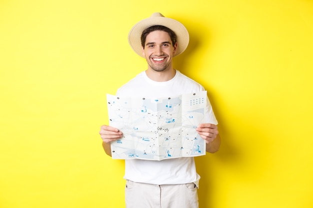 Bezpłatne zdjęcie koncepcja podróży, wakacji i turystyki. uśmiechnięty młody człowiek wybiera się w podróż, trzymając mapę drogową i uśmiechnięty, stojąc na żółtym tle.