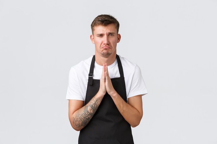 Bezpłatny plik Zdjęcie koncepcja pracowników, sklepów spożywczych i kawiarni. zdenerwowany szlochający barista, kelner w czarnym fartuchu błagający o dzień wolny, trzymający się za ręce w modlitwie i dąsający się, dąsając się do kierownika w kawiarni, białe tło