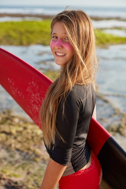 Koncepcja raju surfingowego. Dość młoda surferka z długimi prostymi włosami, nosi deskę surfingową, na twarzy ma odporny na słońce krem cynkowy