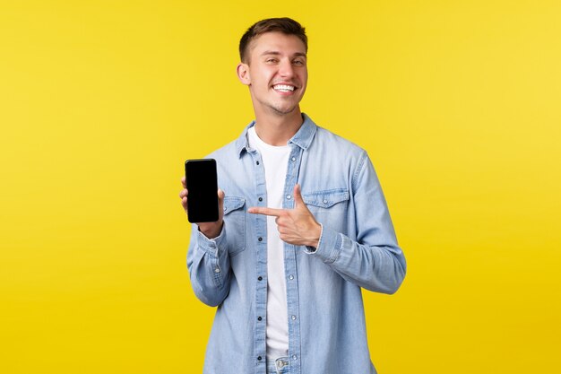 Koncepcja reklamy czasu wolnego, technologii i aplikacji. Dumny szczęśliwy uśmiechnięty blondyn polecający aplikację na smartfona, wskazujący palcem na telefon komórkowy, by chwalić się swoimi zdjęciami, żółte tło.