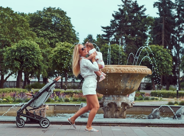 Koncepcja rodziny i ludzi - szczęśliwa matka blondynka ubrana w białą bluzkę i spodenki bawiące się z córeczką w pobliżu fontanny w parku miejskim.