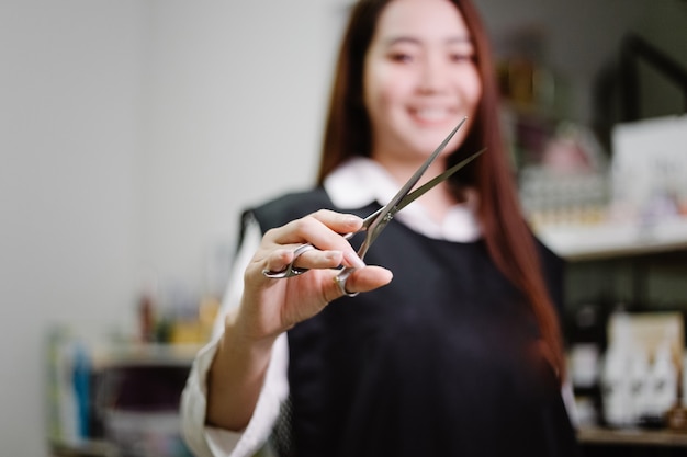 Koncepcja salonu fryzjerskiego ładna fryzjerka pozuje z nożyczkami w dłoni w swoim salonie fryzjerskim w otoczeniu produktów i sprzętu do pielęgnacji włosów.