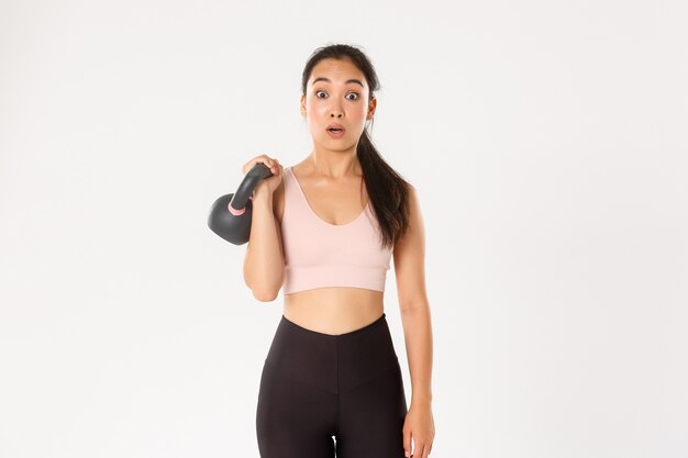 Koncepcja sportu, dobrego samopoczucia i aktywnego stylu życia. Portret ślicznej brunetki azjatyckiej dziewczyny fitness, zapisuje się na zajęcia kulturystyczne na siłowni, zaskoczona ciężarem kettlebell, stojąc na białym tle.