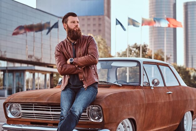 Koncepcja stylu wiejskiego. Brodaty mężczyzna ubrany w brązową skórzaną kurtkę i buty, opierając się na dostrojonym samochodzie retro na parkingu miejskim w pobliżu wieżowca.