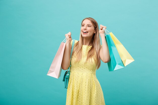 Koncepcja stylu życia: Portret zszokowanej młodej atrakcyjnej kobiety w żółtej letniej dressposing z torby na zakupy i patrząc na kamerę na niebieskim tle.