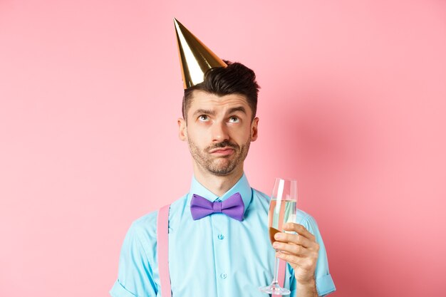 Koncepcja świąt i uroczystości. Zrzędliwy facet w urodzinowym kapeluszu i trzymający kieliszek szampana, patrzący w górę z sceptyczną twarzą, stojący na różowym tle.