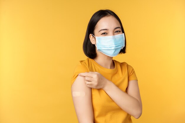 Koncepcja szczepień i pandemii covid19 Uśmiechnięta azjatycka kobieta w medycznej masce na twarz pokazująca ramię z pomocą opaski po szczepieniu z żółtego tła koronawirusa