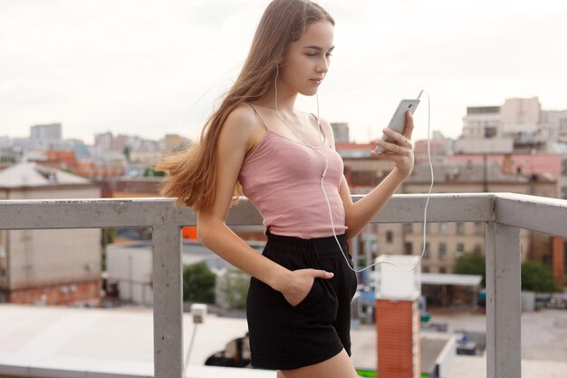 Koncepcja technologii, emocji, ludzi, muzyki, urody, mody i stylu życia - młoda kobieta ze słuchawkami zwisającymi na telefonie komórkowym, gdy spaceruje po miejskiej ulicy, widok z wysokości