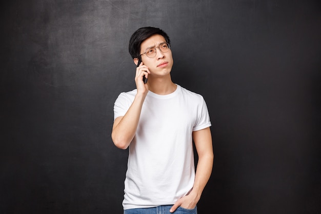 Koncepcja technologii, gadżetów i ludzi. portret rozważny młody azjatykci facet myśleć nad pytaniem, dzwoni przyjaciela, opowiada na telefonie, trzyma smartphone i patrzeje daleko od rozważając, jaka odpowiedź
