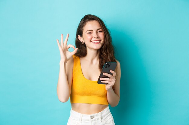 Koncepcja technologii i stylu życia. Piękna dziewczyna ze szczęśliwym uśmiechem, pokazując znak zgody, powiedz tak, trzymając smartfon, stojąc na niebieskim tle.