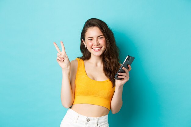 Koncepcja technologii i stylu życia. Piękna wesoła dziewczyna wysyłająca pozytywne wibracje, uśmiechnięta i pokazująca znak pokoju, używająca telefonu komórkowego, stojąca na niebieskim tle.