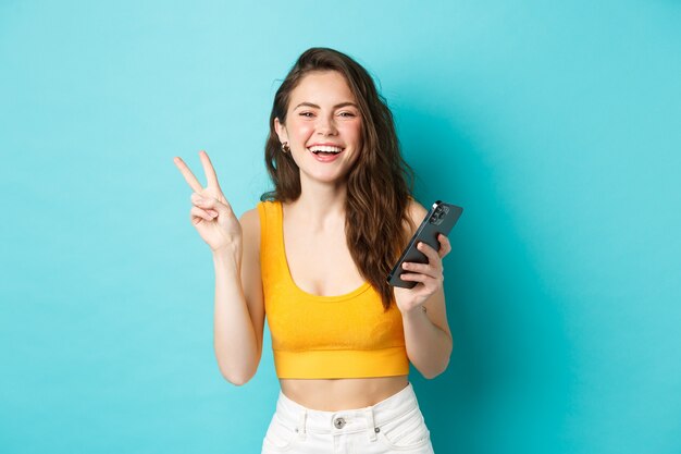 Koncepcja technologii i stylu życia. Szczęśliwa atrakcyjna kobieta śmiejąca się, pokazująca znak V i używająca smartfona, rozmawiająca na telefonie komórkowym, stojąca na niebieskim tle