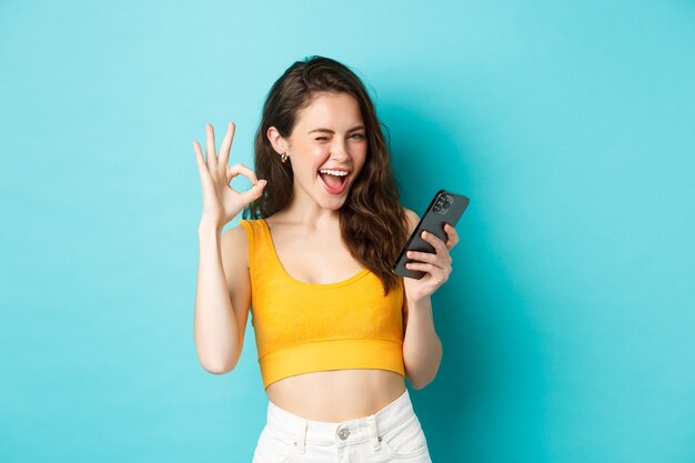 Koncepcja technologii i stylu życia. Wesoła brunetka modelka mówi tak, mrugając i pokazując znak porządku, trzymając telefon komórkowy, stojąc na niebieskim tle.