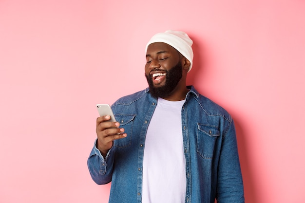 Koncepcja technologii i zakupów online. Szczęśliwy czarny brodaty mężczyzna czytający wiadomość i uśmiechający się, używający smartfona na różowym tle