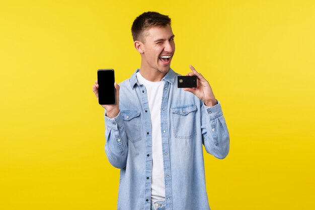 Koncepcja technologii, stylu życia i reklamy. Bezczelny przystojny mężczyzna dokonujący zamówienia online, robiący zakupy w Internecie za pomocą aplikacji mobilnej, pokazujący ekran smartfona i kartę kredytową, żółte tło
