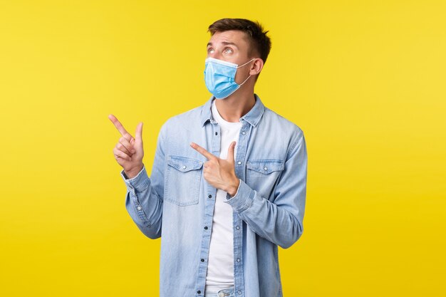 Koncepcja wybuchu pandemii Covid-19, styl życia podczas zdystansowania społecznego koronawirusa. Ciekawy przystojny mężczyzna w masce medycznej, zwróć twarz wskazującą lewy górny róg, czytając znak na żółtym tle.