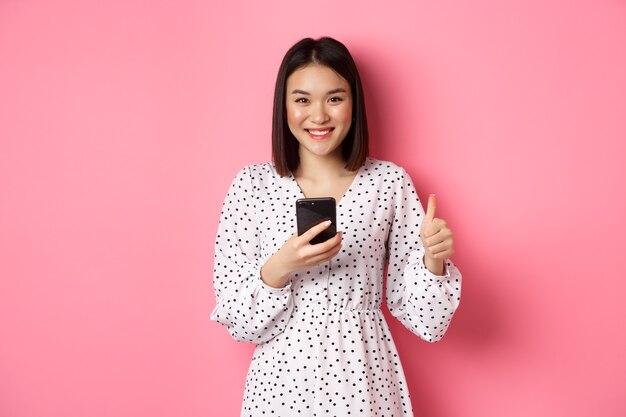 Koncepcja zakupów online i urody. Zadowolona azjatycka klientka pokazując kciuk do góry, dokonując zakupu w internecie na smartfonie, stojąc na różowym tle.
