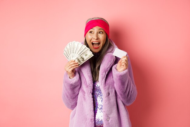 Koncepcja zakupy i moda. Azjatycka starsza kobieta krzyczy szczęśliwa jak zwycięzca, trzymająca pieniądze w dolarach i plastikową kartę kredytową, wyglądająca na podekscytowaną, różowe tło