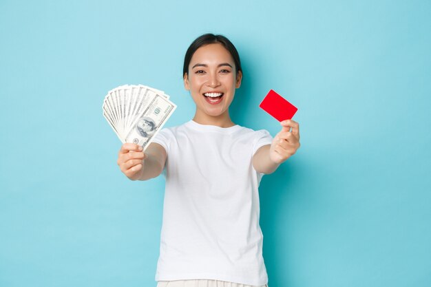 Koncepcja zakupy, pieniądze i finanse. Szczęśliwa i zadowolona uśmiechnięta azjatycka dziewczyna pokazuje dolary w gotówce i karcie kredytowej z dumnym wyrazem, stojąc zadowolona nad jasnoniebieską ścianą.