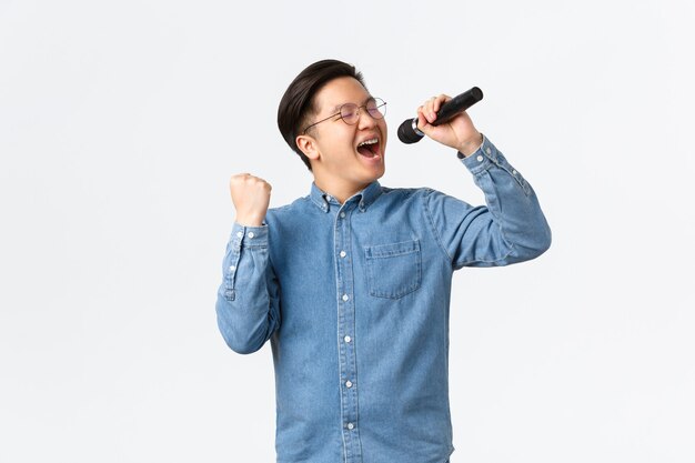 Koncepcja życia, wypoczynku i ludzi. Beztroski, szczęśliwy Azjata, cieszący się śpiewem na karaoke, trzymający mikrofon i pompującą pięścią z zachwytem, występujący na białym tle.