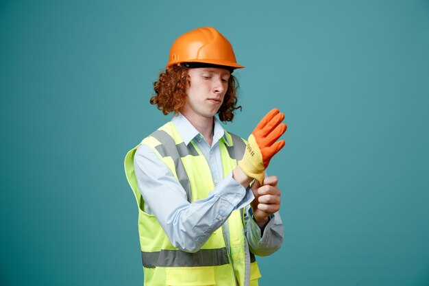 Konstruktor młody człowiek w mundurze budowlanym i kasku ochronnym w rękawiczkach, wyglądający pewnie stojąc na niebieskim tle