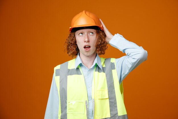 Konstruktor młody człowiek w mundurze budowlanym i kasku patrząc w górę zdziwiony drapiąc się po głowie stojąc na pomarańczowym tle