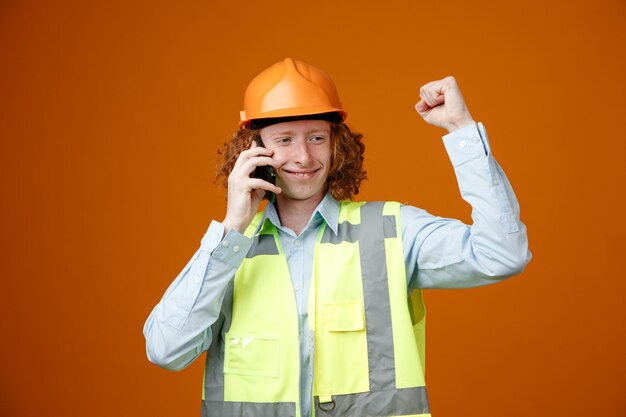 Konstruktor młody człowiek w mundurze budowlanym i kasku rozmawia przez telefon komórkowy szczęśliwy i zadowolony, zaciskając pięść, ciesząc się jego sukcesem, stojąc na pomarańczowym tle