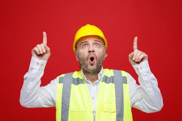 Bezpłatne zdjęcie konstruktor w mundurze budowlanym i kasku ochronnym, patrząc w górę zaskoczony, wskazując palcami wskazującymi w górę, stojąc na czerwonym tle