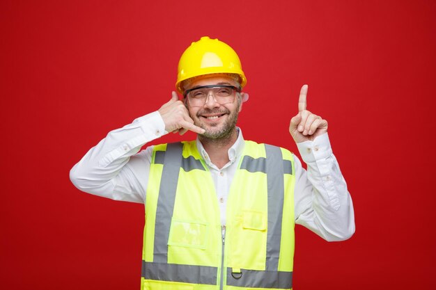 Konstruktor w mundurze budowlanym i kasku ochronnym w okularach ochronnych, patrząc na kamerę, uśmiechając się radośnie, wykonując zadzwoń do mnie gest wskazujący palcem wskazującym w górę, stojąc na czerwonym tle