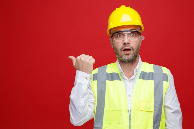 Konstruktor w mundurze budowlanym i kasku ochronnym w okularach ochronnych, patrząc na kamerę zaskoczony, wskazując kciukiem w bok, stojąc na różowym tle