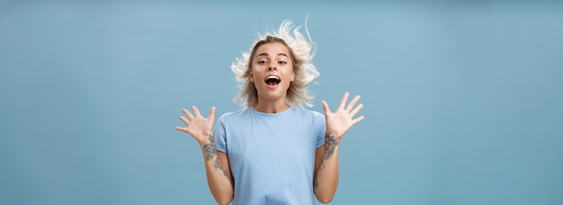 Bezpłatne zdjęcie kreatywna szczęśliwa i figlarna piękna blond dziewczyna z tatuażami na ramionach, unosząc dłonie do góry, otwierając usta