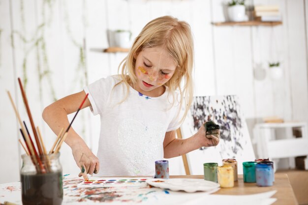 Kreatywne piękne dziecko płci żeńskiej o blond włosach pracuje nad jej zdjęciem w sali sztuki