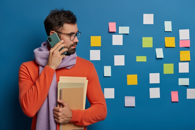 Bezpłatny plik Zdjęcie kreatywny pracownik z brodą dzwoni do kogoś przez telefon komórkowy, nosi okulary, sweter z szalikiem, patrzy na bok na kolorowe notatki przyklejone do niebieskiej ściany