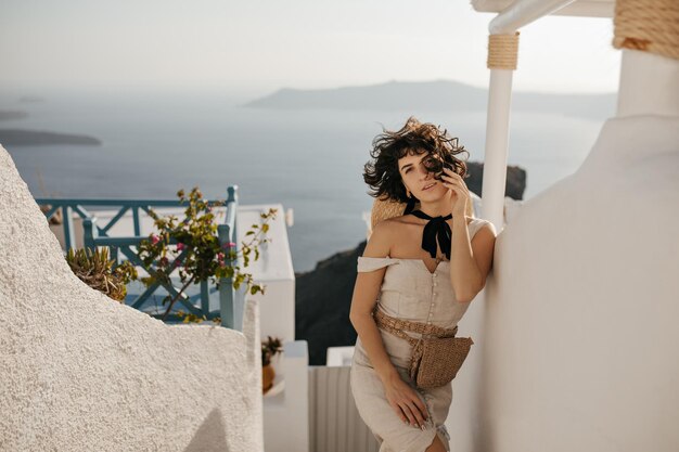Kręcona brunetka dama w beżowym stroju i ze słomianą torbą patrzy do kamery na zewnątrz Ładna młoda kobieta spaceruje po greckim mieście z białymi budynkami i pięknym widokiem na morze