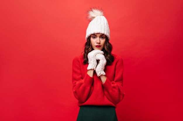 Kręcona kobieta w czerwonym swetrze, dzianinowej czapce i rękawiczkach wygląda z przodu