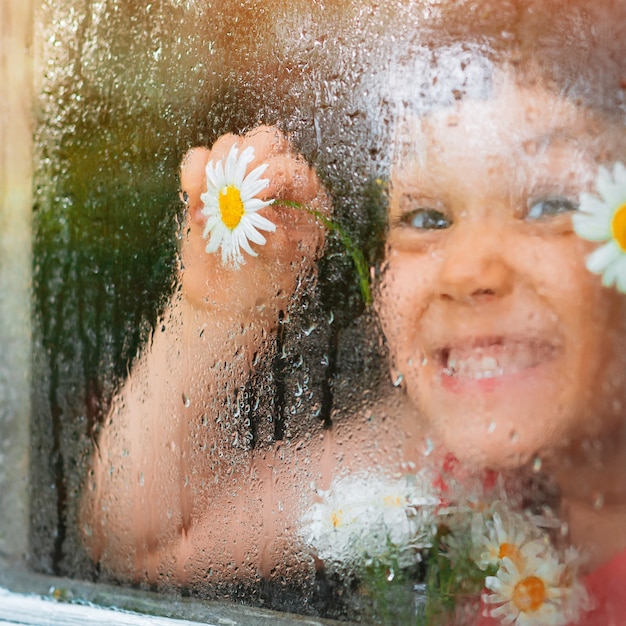 Krople deszczu na szybie wiejskiego okna, oczy z kwiatów rumianku w dłoniach dzieci patrzą na deszcz.
