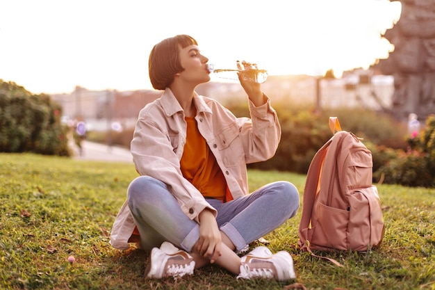 Krótkowłosa kobieta pije wodę na zewnątrz Wesoła młoda stylowa dziewczyna w różowej kurtce i dżinsowych spodniach siedzi na trawie w mieście