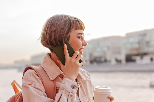 Bezpłatne zdjęcie krótkowłosa kobieta rozmawia przez telefon na zewnątrz optymistyczna dziewczyna w okularach przeciwsłonecznych i beżowa kurtka dżinsowa, trzymając filiżankę herbaty i uśmiechając się na zewnątrz