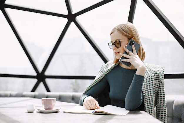 Bezpłatne zdjęcie kryty portret młodej bizneswoman, siedząc w kawiarni i rozmawiając przez telefon. blondynka w okularach przeciwsłonecznych i kurtce