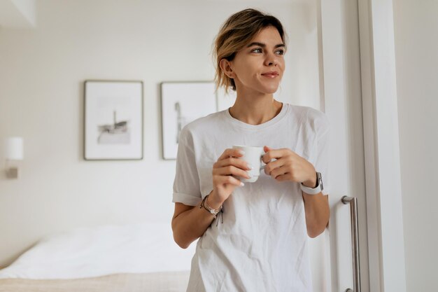Kryty portret młodej kobiety w piżamie trzyma kubek z kawą i patrzy przez okno w nowoczesnym, jasnym mieszkaniu