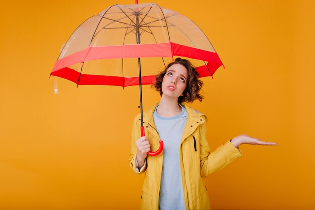 Kryty portret zdenerwowany młodej modelki w jesienny płaszcz przeciwdeszczowy. Zdjęcie smutnej kobiety kręconej stojącej pod modnym parasolem i patrząc w górę.