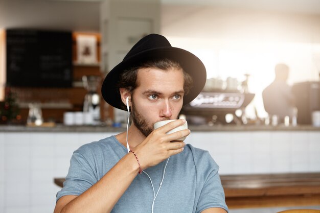 Kryty strzał atrakcyjnego młodego człowieka w stylowym kapeluszu, pije kawę lub herbatę, relaksując się w samej kawiarni, słuchając muzyki online na słuchawkach