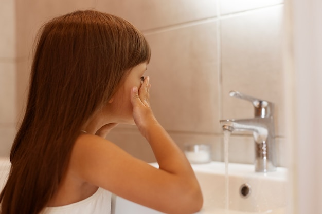 Bezpłatne zdjęcie kryty strzał ciemnowłosy dziecko płci żeńskiej, stojąc w łazience i mycie twarzy po przebudzeniu lub przed pójściem spać, procedury higieniczne w domu.