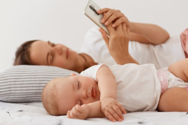 Kryty strzał śpiące dziecko na sobie białą koszulkę, matka leżąca w pobliżu córki i trzymająca telefon komórkowy, kobieta za pomocą smartfona do przeglądania Internetu lub pisania wiadomości.